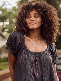 A woman with voluminous curly brown hair wearing a dark charcoal gray short-sleeve top with colorful floral embroidery, photographed outdoors with a blurred background.