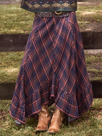 A high-waisted, flared plaid skirt in navy blue with red and orange stripes, styled with a western belt and brown cowboy boots, shown outdoors in front of a wooden fence.