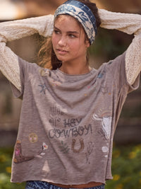 A woman wearing a distressed taupe-brown graphic t-shirt with 'HEY COWBOY!' text and cowboy boot illustrations, styled with a blue patterned headband and a cream knit cardigan.