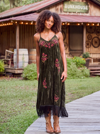 A woman stands on a wooden boardwalk in front of a rustic wooden bungalow, wearing a dark olive green velvet midi dress with pink floral embroidery and black fringe, paired with brown cowboy boots.