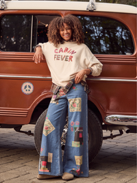 A woman stands in front of a vintage brown van, wearing a cream sweatshirt with 'Cabin Fever' printed in red and blue jeans covered in colorful patchwork fabric squares.