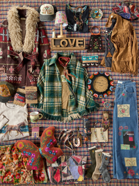 A styled flat-lay collection of bohemian, western, and vintage-inspired fashion items arranged on a red and blue plaid blanket. The collection includes a green and blue plaid fringed cardigan, a brown faux-fur collar, a fringed tan vest, a patchwork denim skirt, embroidered boots, a patterned hat, a small patterned bag, a wooden 'LOVE' sign, and various other bohemian accessories.