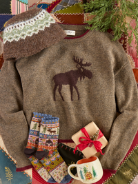 A cozy flat-lay of a brown knit sweater with a dark moose motif on the chest, styled with a matching knit hat, patterned socks, a small wrapped gift, a mug, and evergreen branches on a patchwork quilt background.