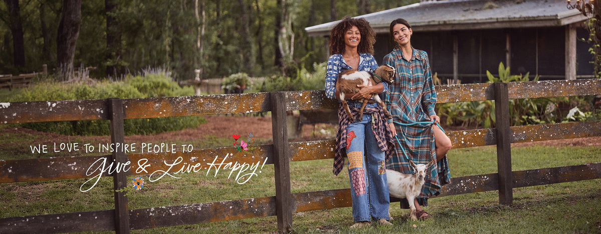 Two women in patchwork jeans and plaid, smiling at a wooden fence with two baby goats, with the text “We love to inspire people to Give & Live Happy!”