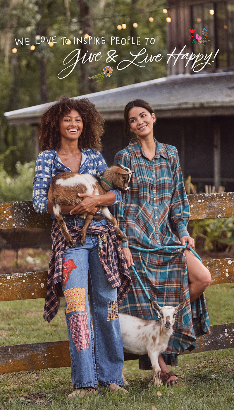 Two women in patchwork jeans and plaid, smiling at a wooden fence with two baby goats, with the text “We love to inspire people to Give & Live Happy!”