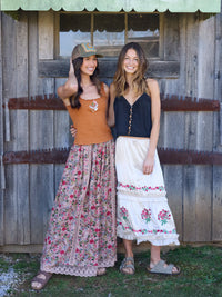 Two women standing in front of a wooden building, wearing casual outfits with floral patterns.