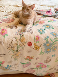 An orange tabby cat lies on a cream-colored blanket printed with colorful flowers, birds, and small animals, photographed in a real indoor setting.