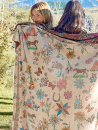 A large woven blanket with a western-themed print of horses, flowers, butterflies, and barns is draped over two women outdoors in a sunny field.