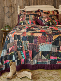 A colorful patchwork quilted bedspread with matching pillows and fringed edges, styled on a bed in a rustic bedroom setting with a wooden headboard, wall decor, boots, and a patterned rug.