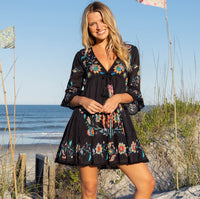 Woman in a black floral dress standing on a beach with ocean and sky in the background