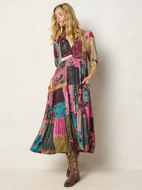 A woman modeling a long patchwork midi dress with a multicolor floral and geometric print, featuring a button-down front, three-quarter sleeves, and a matching fabric belt. She is wearing brown cowboy boots against a plain off-white studio background.