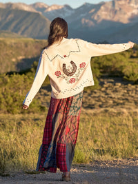 A woman stands outdoors in a grassy mountain landscape, viewed from behind, wearing a cream embroidered cardigan with a large horseshoe and rose design on the back, paired with a long patchwork skirt.