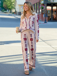 A woman walking outdoors on a sunny street wearing a matching long-sleeve button-front top and wide-leg pants set in a light cream color with dark red floral embroidery. She is holding a paper coffee cup.