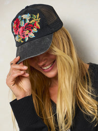 A woman wearing a black trucker hat with colorful floral beadwork on the front panel, holding the brim with one hand against a plain light background.