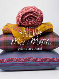 A stack of folded and rolled sheets in assorted prints and patterns, including floral and plaid designs, with overlaid text reading 'NEW Mix + Match prints are here!' on a light gray background.