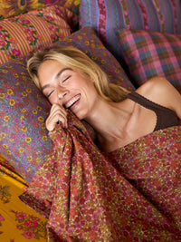 A woman lying down smiling and laughing, wrapped in a rust-red floral patterned blanket, with matching patterned pillows behind her.