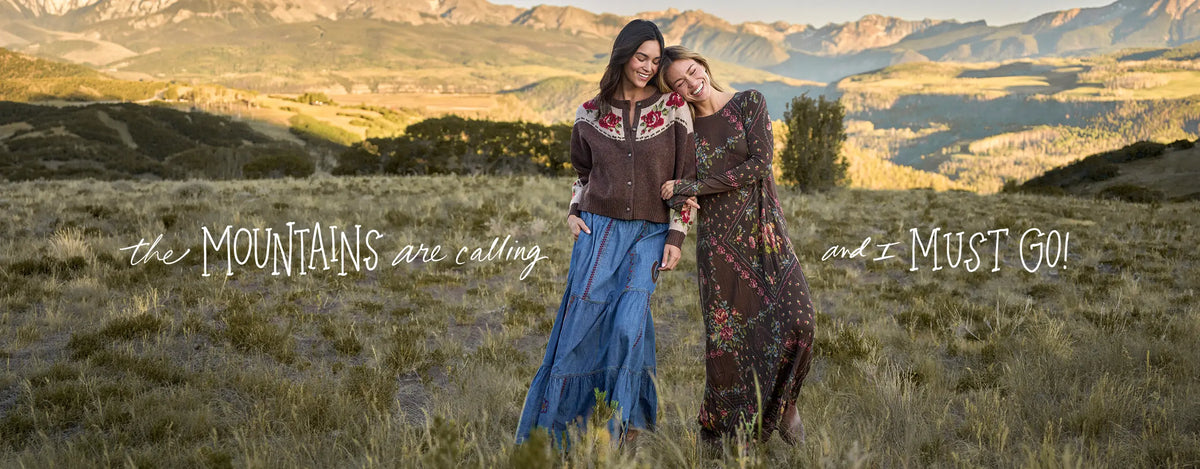 Two women standing in a grassy mountain landscape at golden hour, wearing long patterned and embroidered dresses. White script text overlays read 'the MOUNTAINS are calling and I MUST GO!'.