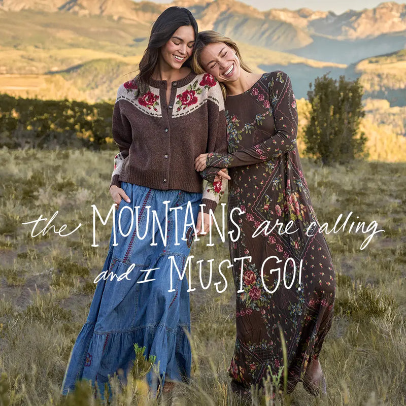 Two women standing outdoors in a grassy mountain landscape, smiling and embracing. One wears a brown embroidered cardigan with a blue skirt, and the other wears a dark floral long-sleeve dress.