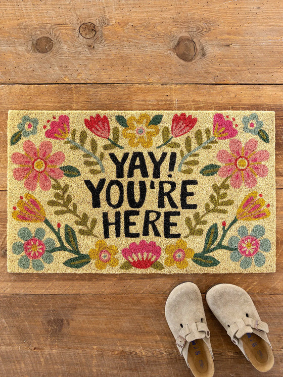 Decorative doormat with floral design and 'YAY! YOU'RE HERE' text on a wooden floor.