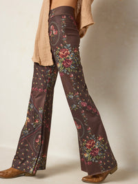 A person wearing brown flared pants with a floral and paisley print, styled with a beige cardigan and brown boots against a neutral studio background.
