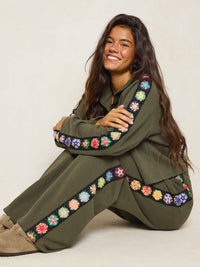 A woman sitting on the floor wearing an olive green matching lounge set with colorful floral embroidered trim along the sleeves and legs, posed against a plain light beige studio background.