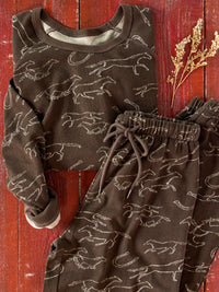 A two-piece children's pajama set featuring a long-sleeve top and matching pants in dark brown with a white line-art pattern of running dogs, styled on a rustic red wooden background with dried flowers.