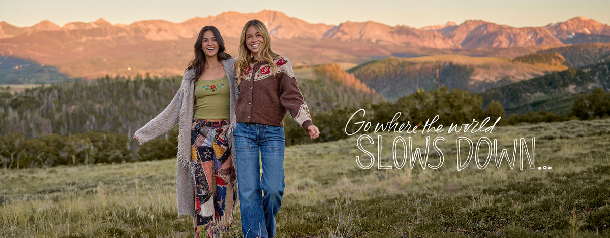 Two women walking hand in hand in a grassy field with mountains in the background at sunset. One wears a green top, patterned skirt, and beige cardigan, and the other wears a brown floral jacket and blue jeans.