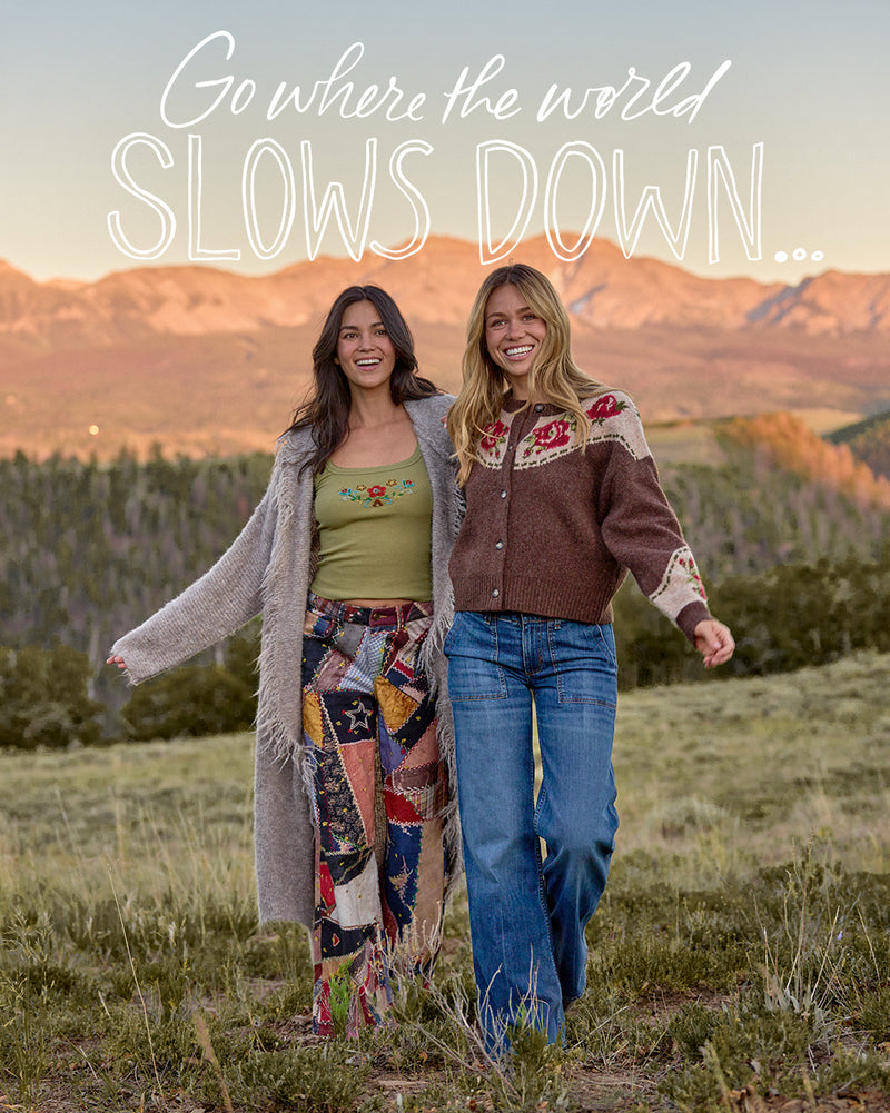 Two women walking outdoors in a grassy mountain landscape at sunset, wearing bohemian-style clothing. Large white text at the top reads, 'Go where the world SLOWS DOWN.