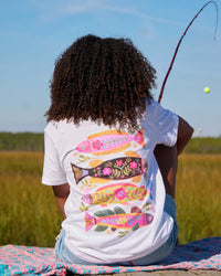 Person wearing a white t-shirt with colorful fish design, sitting in a field with a fishing rod.