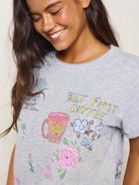 A woman wearing a light gray heather graphic t-shirt with a colorful print of a coffee mug, flowers, and the text 'BUT FIRST COFFEE'.