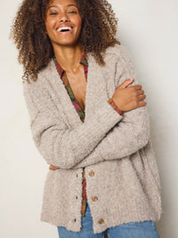 A woman with curly hair smiling and crossing her arms, wearing a fuzzy light beige button-front cardigan over a patterned top and blue jeans, against a plain light background.