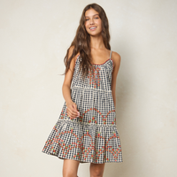 A woman modeling a sleeveless tiered dress with a black and white gingham pattern and orange floral accents, set against a plain beige studio background.