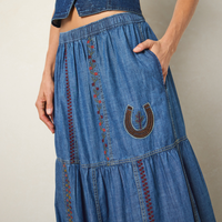 A close-up of a person wearing a blue denim tiered maxi skirt with embroidered details and a brown horseshoe patch on the front pocket.