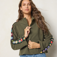 A woman wearing an olive green quarter-zip fleece jacket with colorful floral embroidery along the sleeves, paired with blue jeans, set against a plain light beige background.