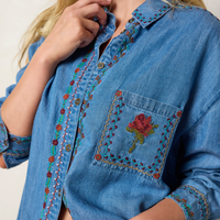 A close-up of a person wearing a blue denim button-up shirt with colorful floral embroidery running down the front placket and on the chest pocket, which features a red rose motif.